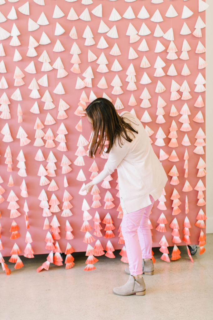 A backdrop made from colored coffee filters folded into cone shapes and strung together in strands. The coffee filters are dyed in various coral shades and are arranged in an ombre pattern.