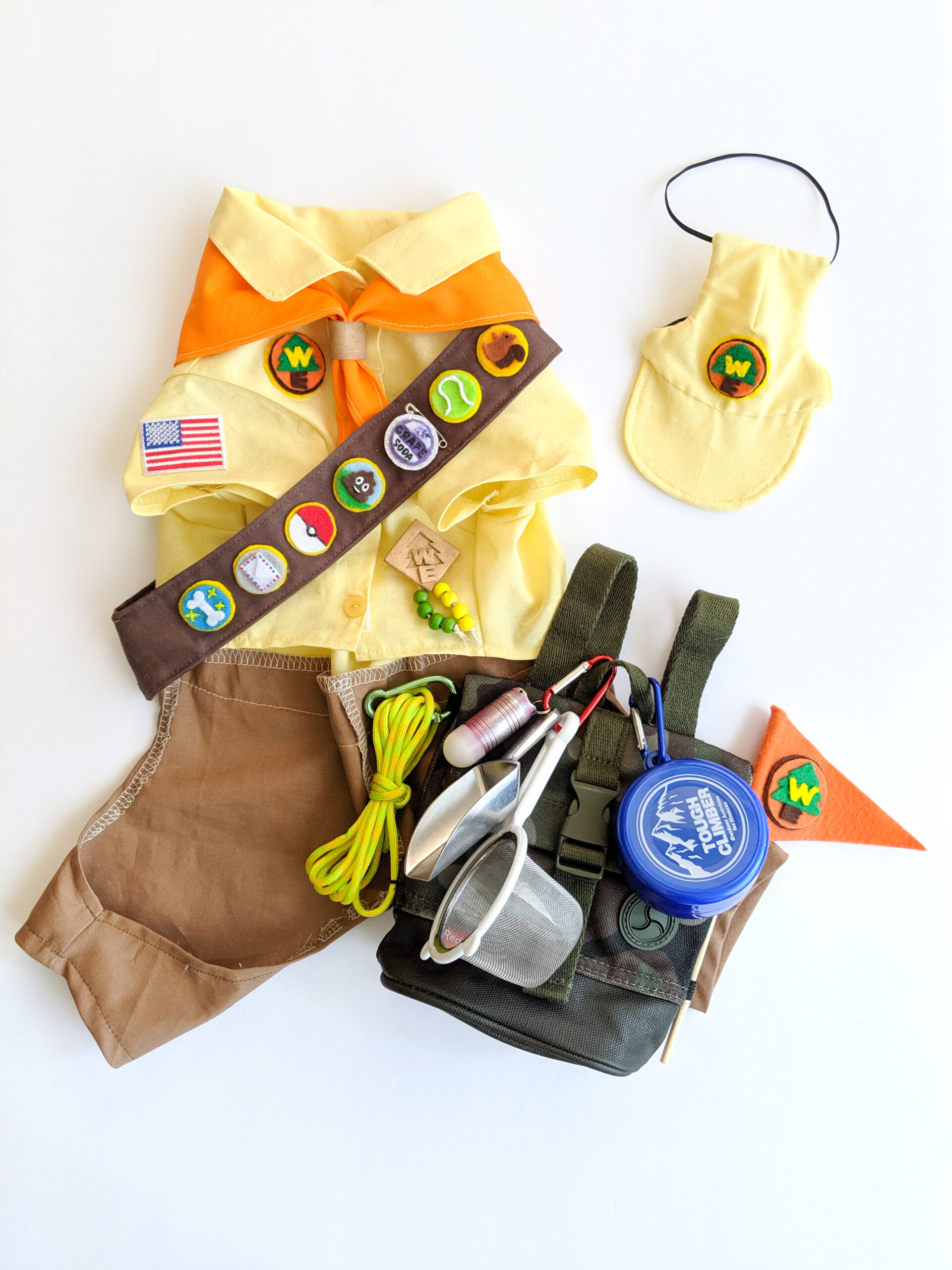 Handmade Wilderness Explorer dog costume worn by Russell in Pixar Up. The top of the costume is a yellow shirt with the Wilderness Explorer logo, an orange bandana, and a sash with handmade merit badges. To the right of the costume is a handmade yellow hat with the WE logo. There's also a backpack with attached tools and a Wilderness Explorer flag.