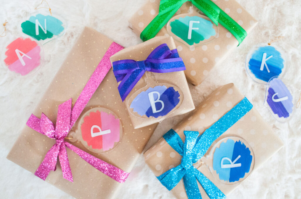 Finished holiday gift wrap hack - painted ornament gift tags. Four brown packages are wrapped in brown paper with pink, purple, green and blue ribbon. Attached are the painted gift tags made by Blaire from Freshly Fuji.