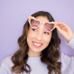 Blaire is wearing her DIY rhinestone sunglasses. She's lifting them over her eyes and smiling. She is wearing a lavender sweater and is in front of a lavender backdrop.