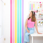 Blaire, a petite Asian American woman with brown hair is dressed in a bright pink top and light blue pants. She is sitting on her white craft room desk and looking up at her rainbow stripe wall.