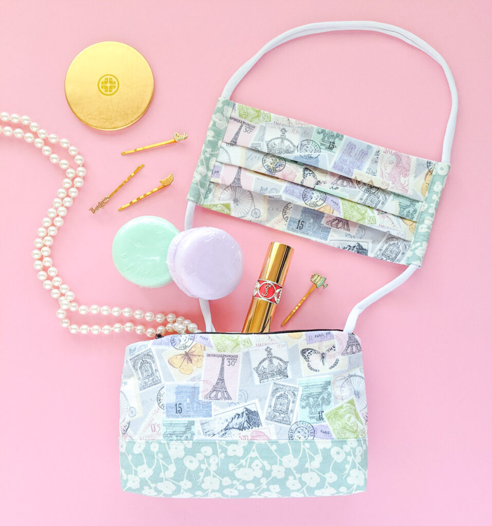 Cosmetic pouch with pastel stamp print on the bottom of the photo with matching face mask coming out of it. Other items leaving the pouch include a pearl necklace, hair clips, lip stick, soap and a gold mirror.