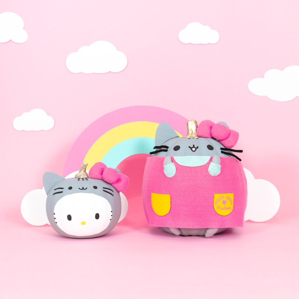 Two pumpkins decorated to look like Pusheen the cat and Hello Kitty. Hello Kitty is wearing a gray Pusheen headpiece and Pusheen is wearing a pink Hello Kitty dress. They both have big pink felt bows on their heads. Behind them is pink backdrop with a pastel rainbow and clouds.