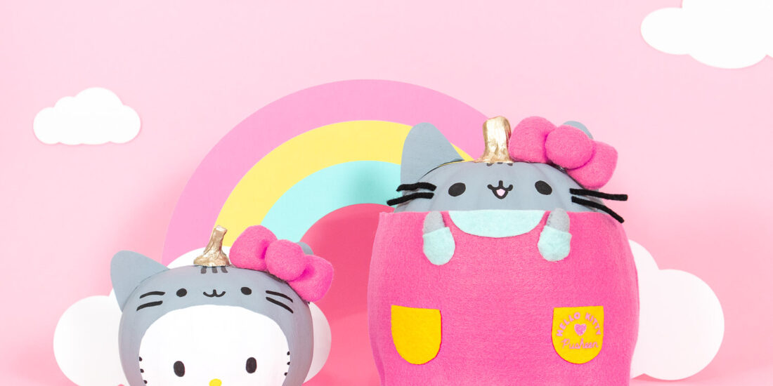 Two pumpkins decorated to look like Pusheen the cat and Hello Kitty. Hello Kitty is wearing a gray Pusheen headpiece and Pusheen is wearing a pink Hello Kitty dress. They both have big pink felt bows on their heads. Behind them is pink backdrop with a pastel rainbow and clouds.