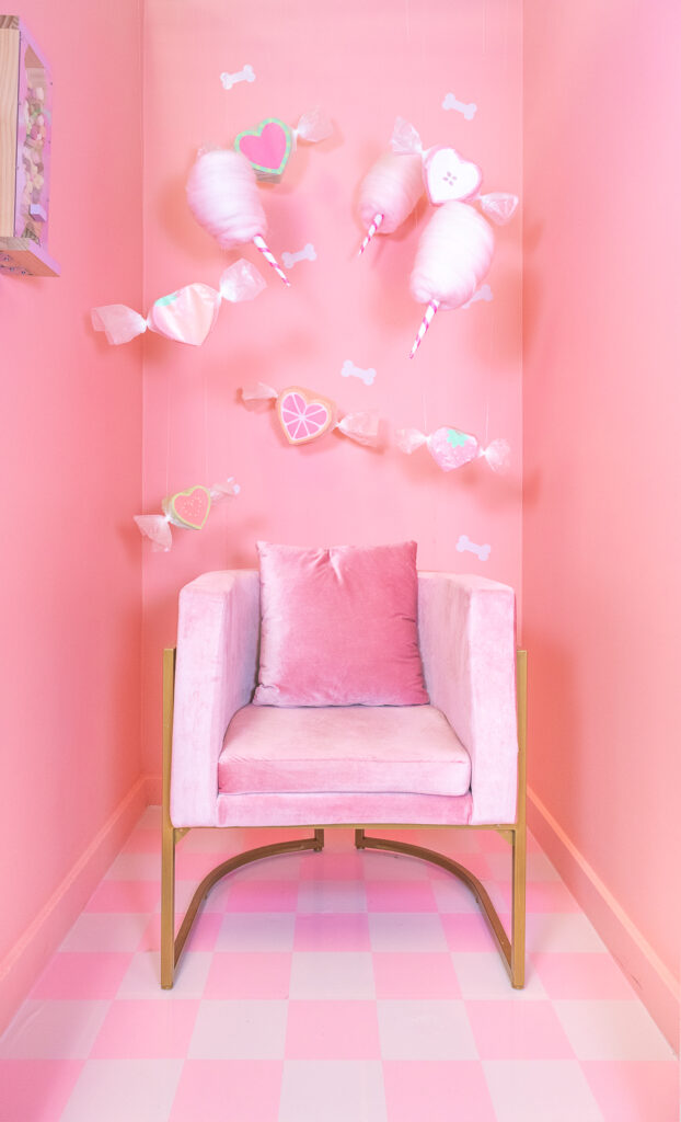 A photo booth for dogs decorated with a candy shop theme. The walls are painted pink and have white dog bone shapes and the floors have pink vinyl applied in a checkerboard pattern. There are also giant handmade cotton candy and salt water taffy decorations suspended from the ceiling. In the center is a modern pink velvet chair with a matching throw pillow.