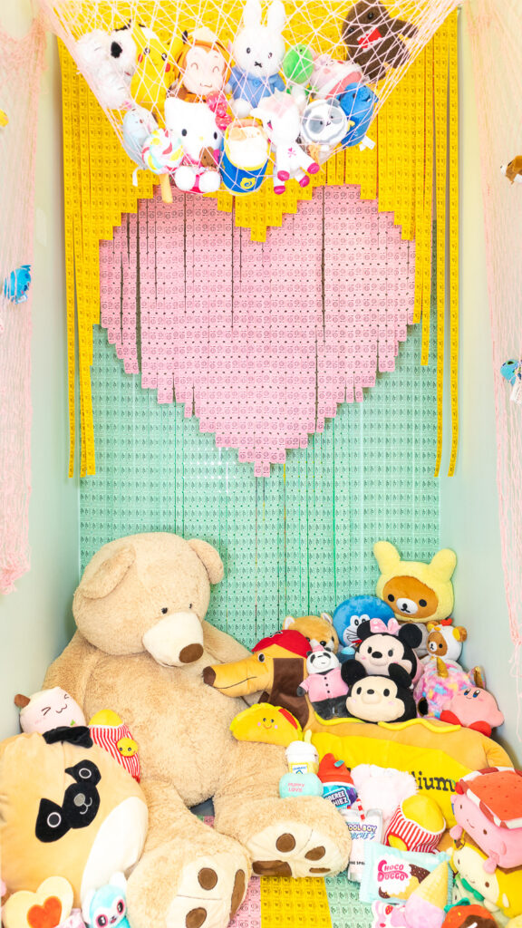 A photo booth for dogs decorated to look like a prize room at an arcade or carnival. The back wall features a layered ticket backdrop with yellow, pink, and green tickets forming a heart shape. There are also tickets in a stripe pattern on the floor. A pink net hangs at the top of the room and is filled with cute stuffed animals and plushies. The bottom of the room is covered in more plush toys of all sizes including a giant teddy bear.