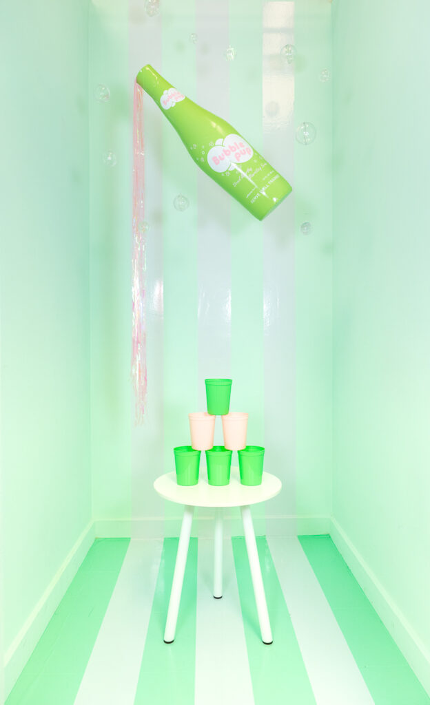 A photo booth for dogs decorated with a soda shop theme. The walls and floors are a light mint green. There are three, large white stripes running from the walls down onto the floor. Hanging from the ceiling is a giant, lime green inflatable soda bottle labeled "Bubble Pup" with iridescent streamers pouring out to look like soda. There are also iridescent bubbles floating around the room. In the center of the room is a small, round, white table with pink and green cups stacked to form a pyramid.