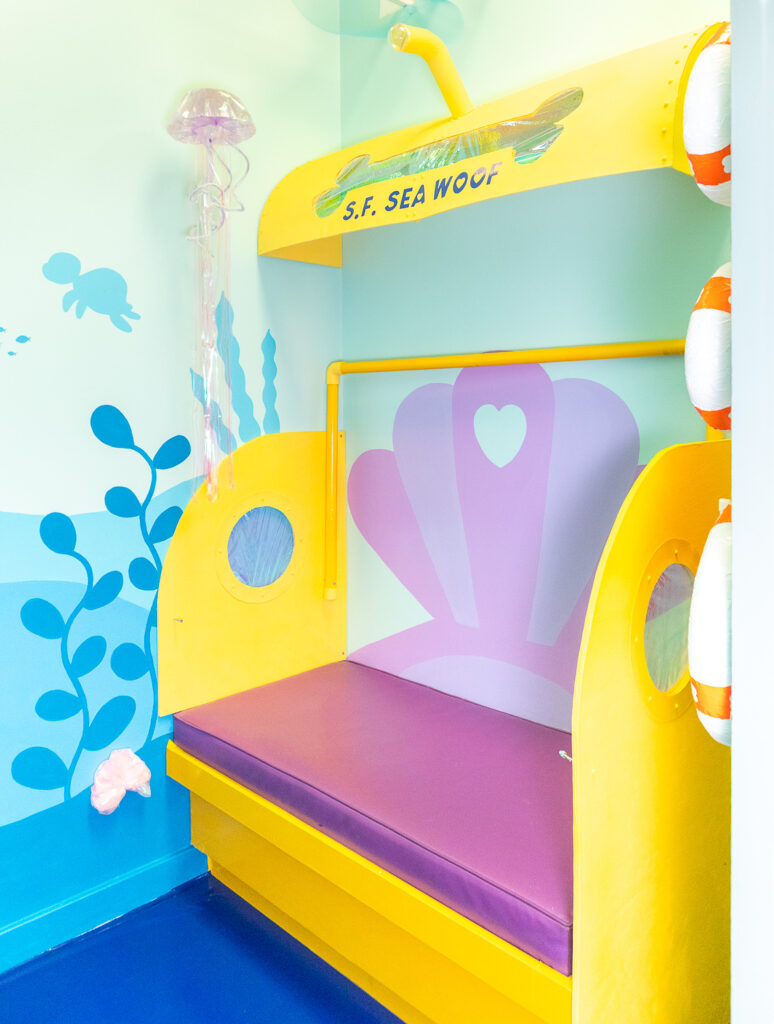 A photo booth for dogs decorated to look like a submarine water ride. The walls are painted with a colorful, under the sea mural featuring seaweed and turtles. The highlight of this room is a giant yellow submarine complete with lookout windows, a safety bar, and a purple seashell bench seat. There is also a light purple jellyfish suspended from the ceiling and life saver props hanging from the walls.