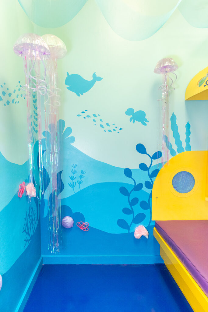 A photo booth for dogs decorated to look like a submarine water ride. The walls are painted with an under the sea mural featuring seaweed, turtles, and fish in various shades of blue. Off to the right is a giant yellow submarine with a purple bench seat. There are also light purple jellyfish suspended from the ceiling and turquoise netting draped from the ceiling.