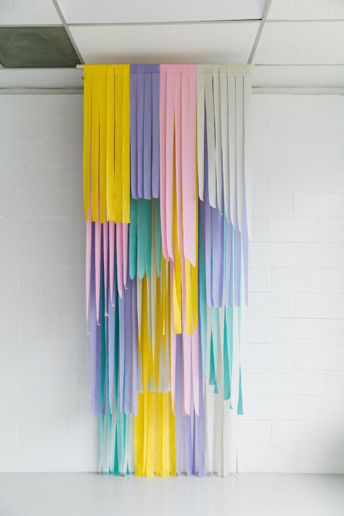 A colorful fringe backdrop featuring overlapping strips of fabric in pastel colors. The backdrop is hanging from the ceiling in a white photo studio.