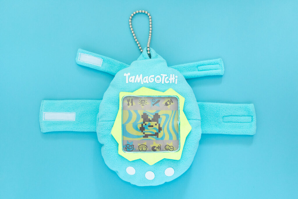 A light blue Tamagotchi dog costume on a blue backdrop. The costume is made from fleece and has a fluorescent green screen with lit screen. There is also a white "Tamagotchi" logo, white buttons, and a ball chain at the top.