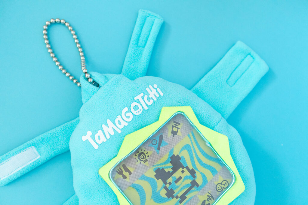 A close-up of a light blue Tamagotchi dog costume on a blue backdrop. The costume is made from fleece and has a fluorescent green screen with lit screen. There is also a white "Tamagotchi" logo and a ball chain at the top.