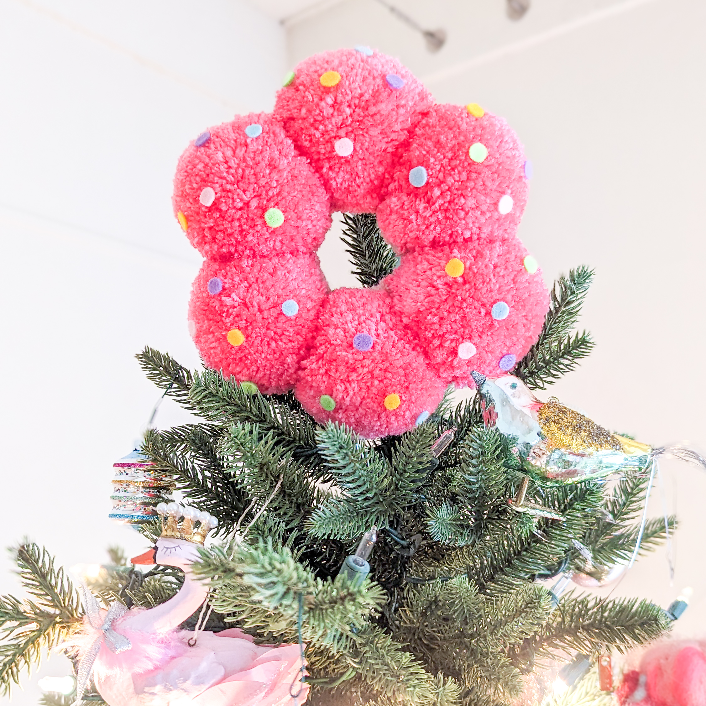 A hot pink tree topper designed to look like a mochi donut. It's decorated with round, felt sprinkles and placed on top of an artificial Christmas tree.