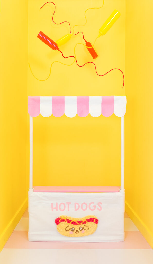 A photo booth room for dogs decorated with a hot dog theme. The room is painted in bright yellow and the floors have a large pink and white stripe pattern. In the center of the room is a hot dog stand labeled "hot dogs" with a handmade punch needle hot dog and a pink and white striped, scalloped awning. Above the hot dog stand are suspended red ketchup and yellow mustard squirt bottles with yarn squiggles that look like ketchup and mustard.