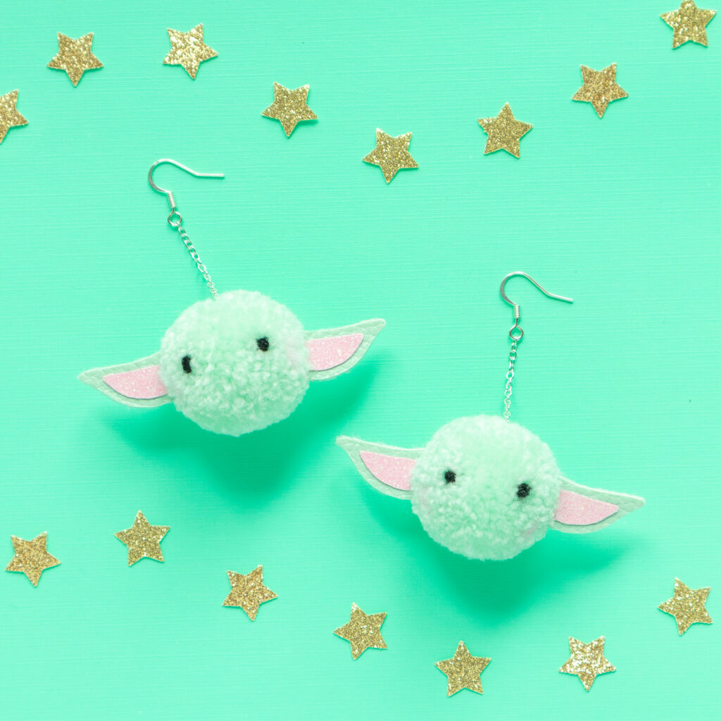 Round, mint green pom pom earrings that look like Grogu from Star Wars, The Mandalorian. The earrings are placed on a mint green background and are surrounded by gold glitter star cut outs. 