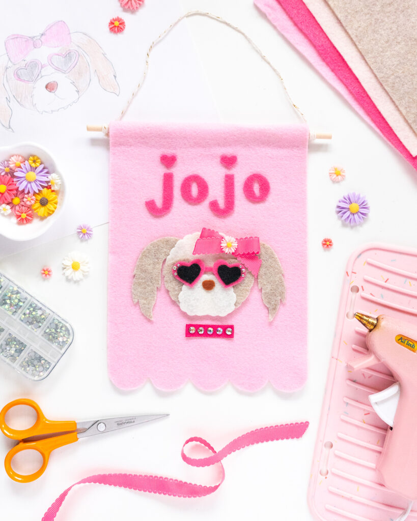 A light pink felt banner with a portrait of a mini Aussie-doodle mix cut from felt. The dog is wearing hot pink sunglasses and a bright pink hair blow. Above the dog's head is "JoJo" cut out from hot pink felt.