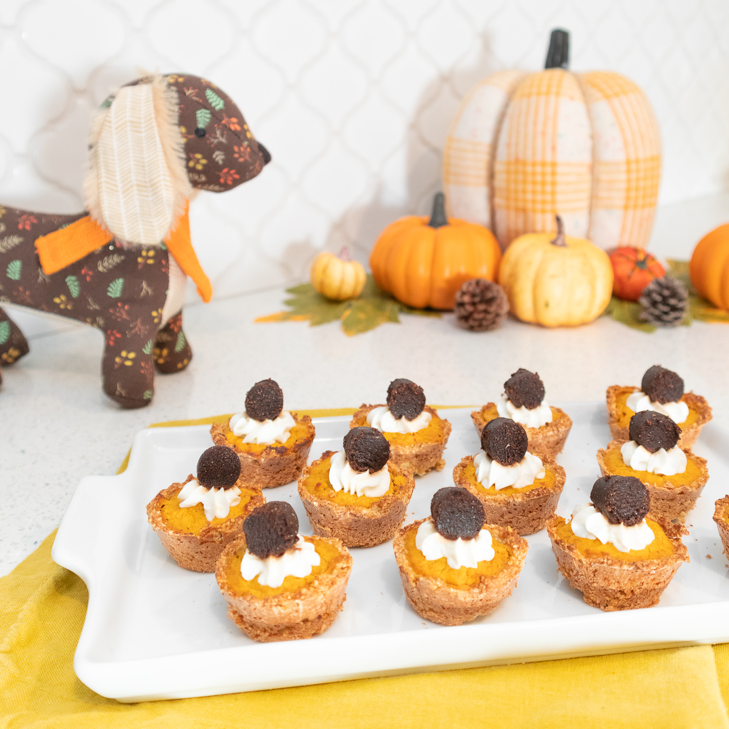 Mini pumpkin pie bites, topped with whipped cream and treats are displayed on a white tray. The tray sits on a mustard yellow napkin. Behind the tray is an assortment of artificial pumpkins and a brown plush dog with a floral pattern all over it.