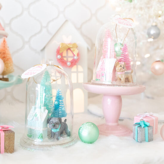 A close up of two DIY pet snow globe ornaments. The one on the left features a small gray dog, a light blue house and mint and blue bottle brush trees. The one on the right is propped on a pink mini cake stand and features a tan Yorkie dog, a pink house, and a pink bottle brush trees.