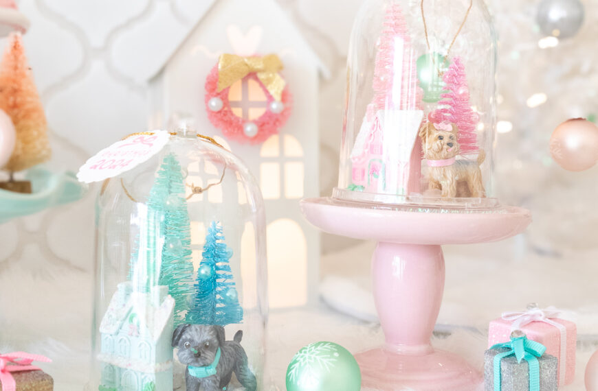 A close up of two DIY pet snow globe ornaments. The one on the left features a small gray dog, a light blue house and mint and blue bottle brush trees. The one on the right is propped on a pink mini cake stand and features a tan Yorkie dog, a pink house, and a pink bottle brush trees.